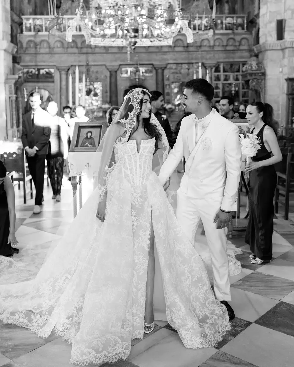 Black and white photo of a bride in a lace gown and a groom in a white suit holding hands in church