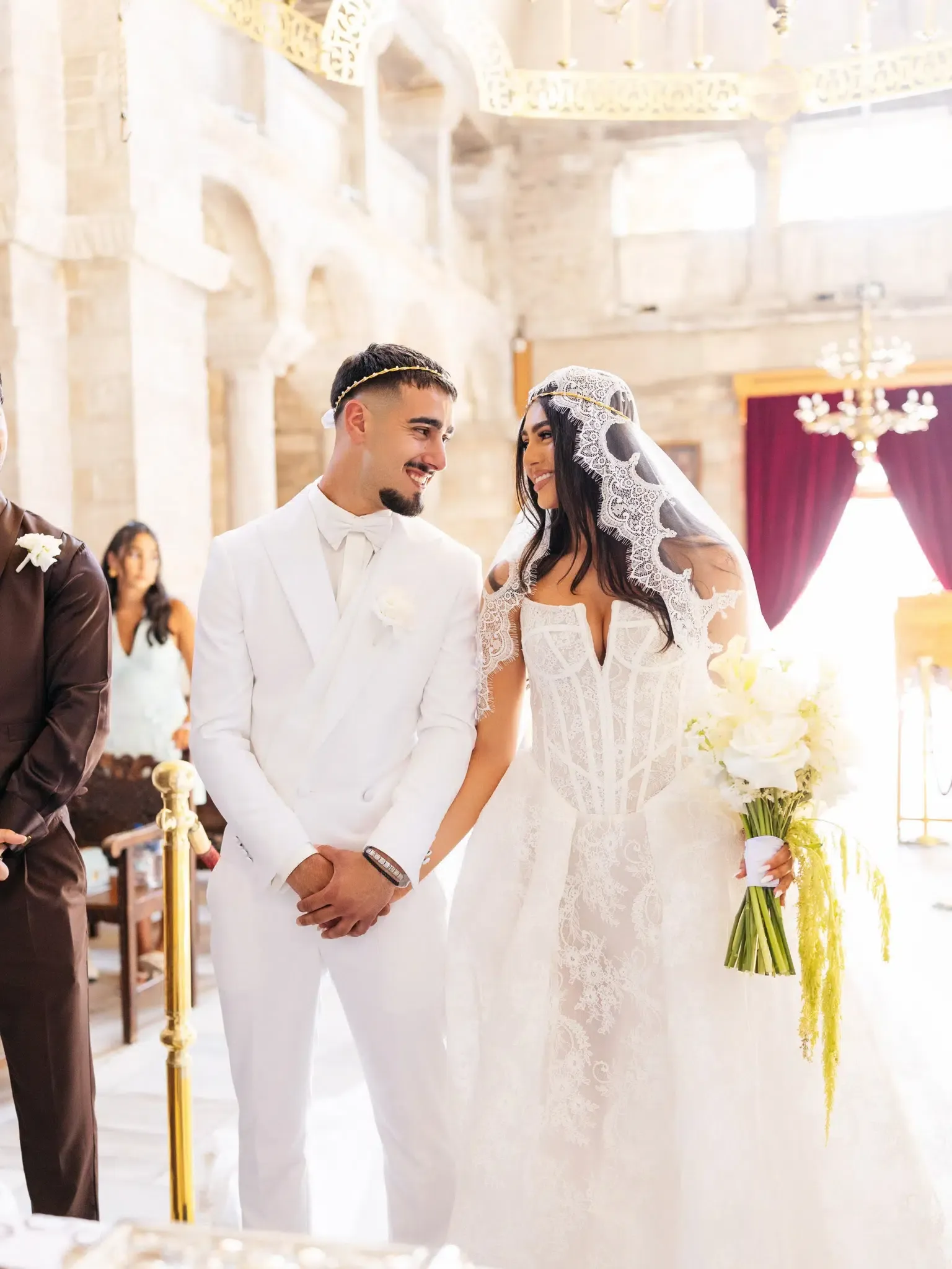 Bride and groom wearing traditional gold wedding crowns, smiling at each other during a church ceremony
