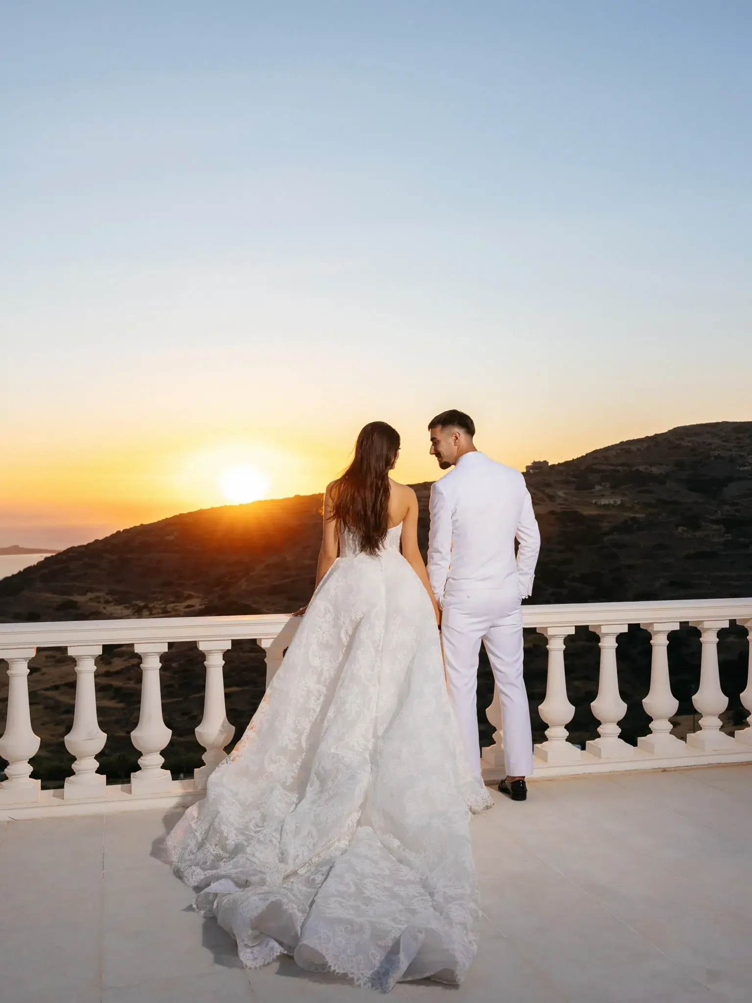 Bride and groom standing on a white stone terrace looking out over hills and the sea at sunset