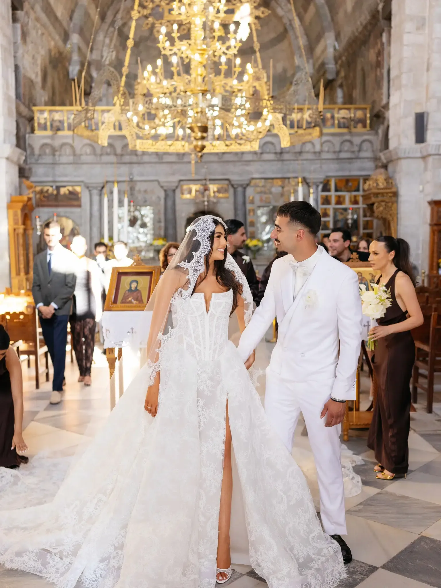 Bride and groom in white holding hands and smiling as they walk the aisle of an ornate church