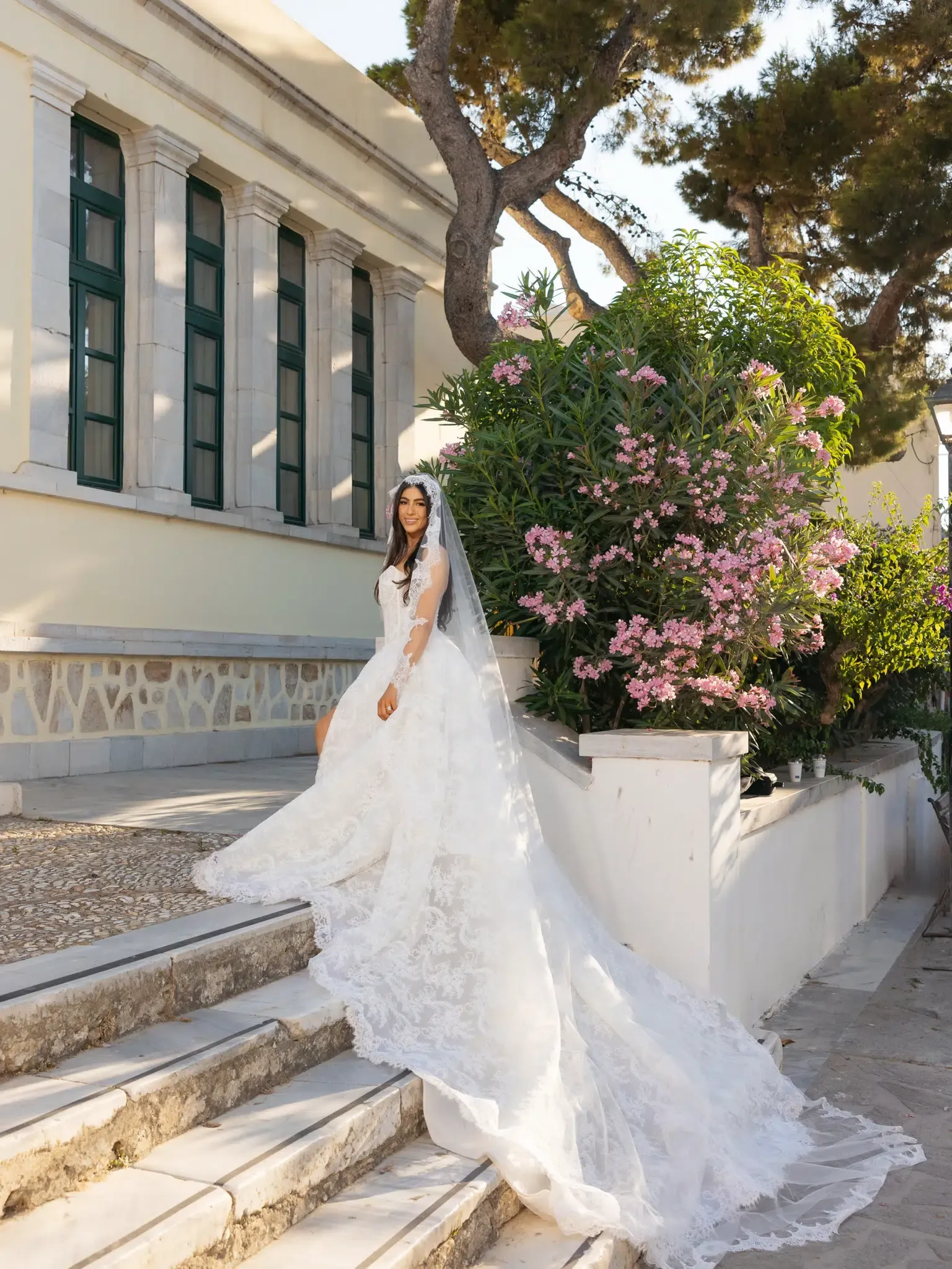 Bride in a long-sleeved lace gown and veil posing on stone steps beside a flowering bush
