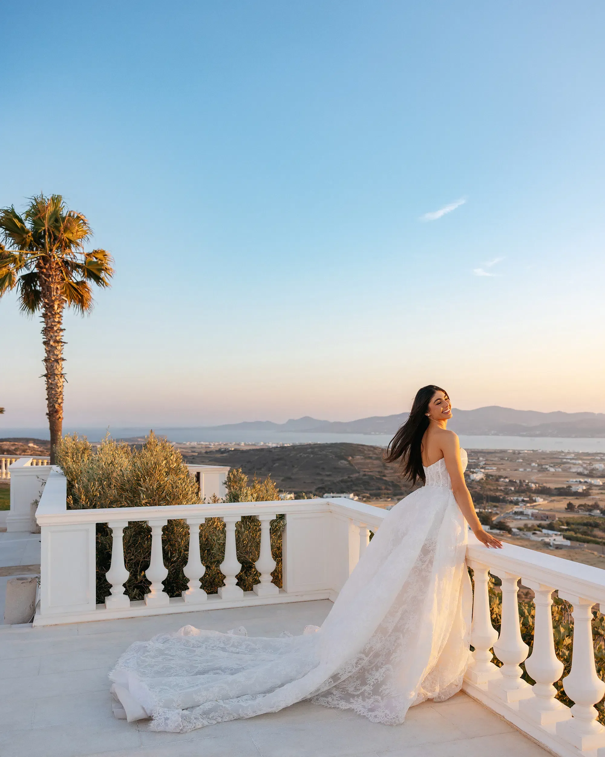 Bride in a strapless lace gown looking back over her shoulder from a balcony overlooking the coast at sunset