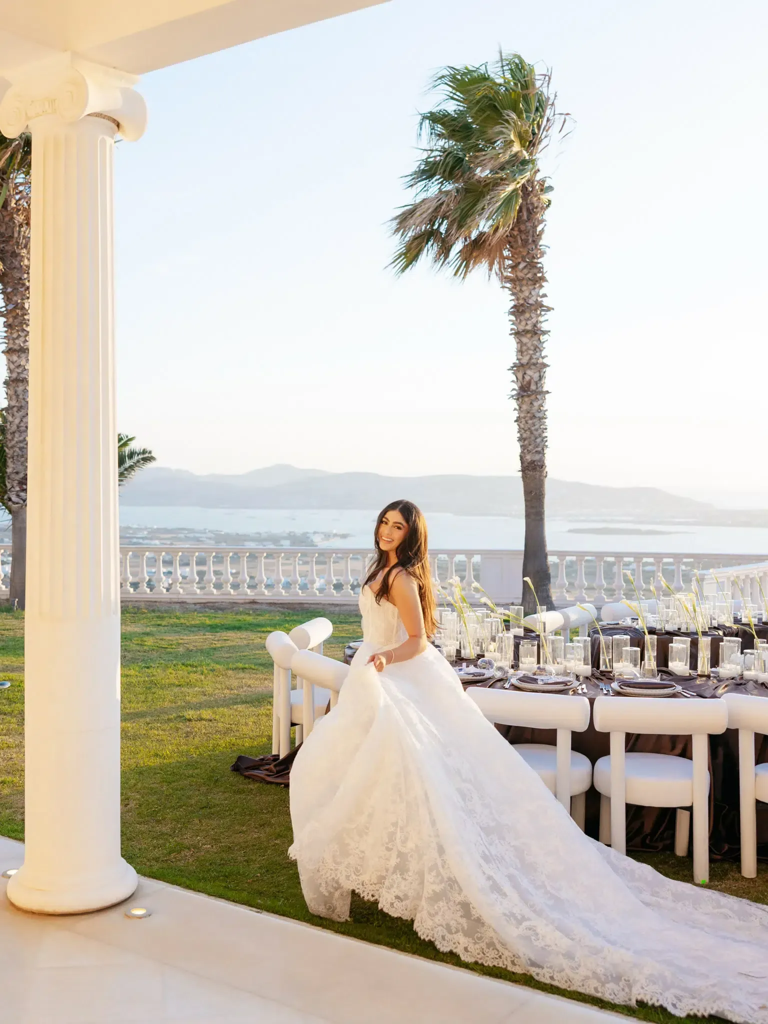 Bride in a strapless lace gown at an outdoor reception with a candlelit table, palm trees and a coastal view