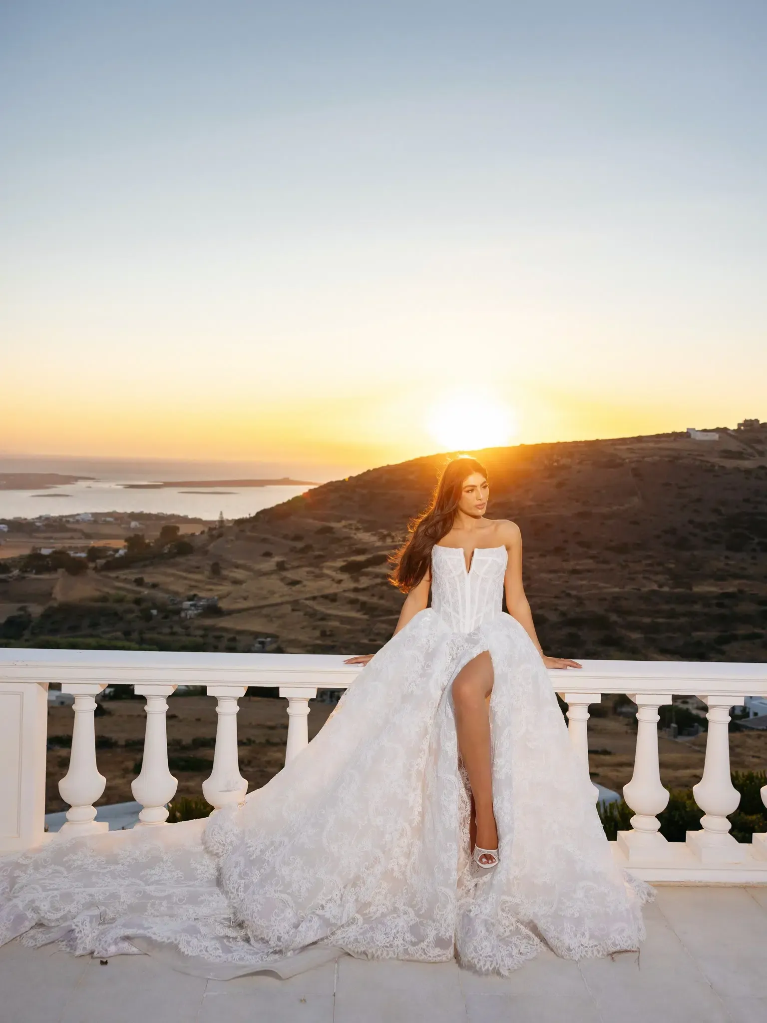 Bride in a strapless lace gown sitting on a white balcony at sunset with a coastal landscape behind her