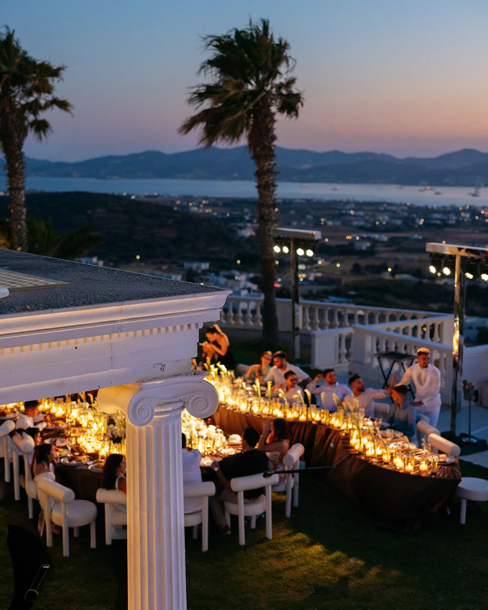 Guests celebrating around a winding candlelit dinner table on a terrace overlooking the coast at dusk