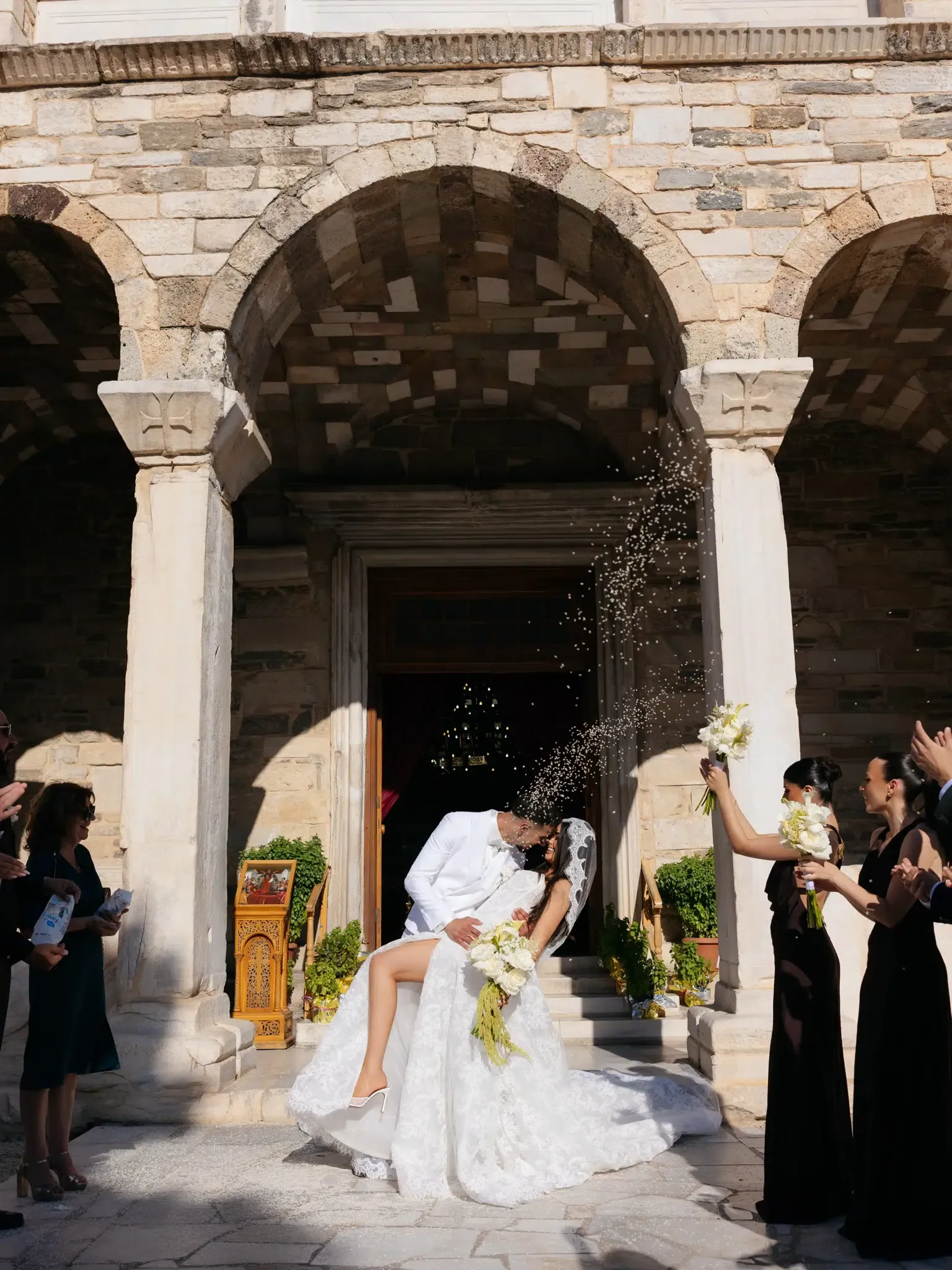 Groom in a white suit dipping the bride for a kiss outside a stone church as guests throw rice