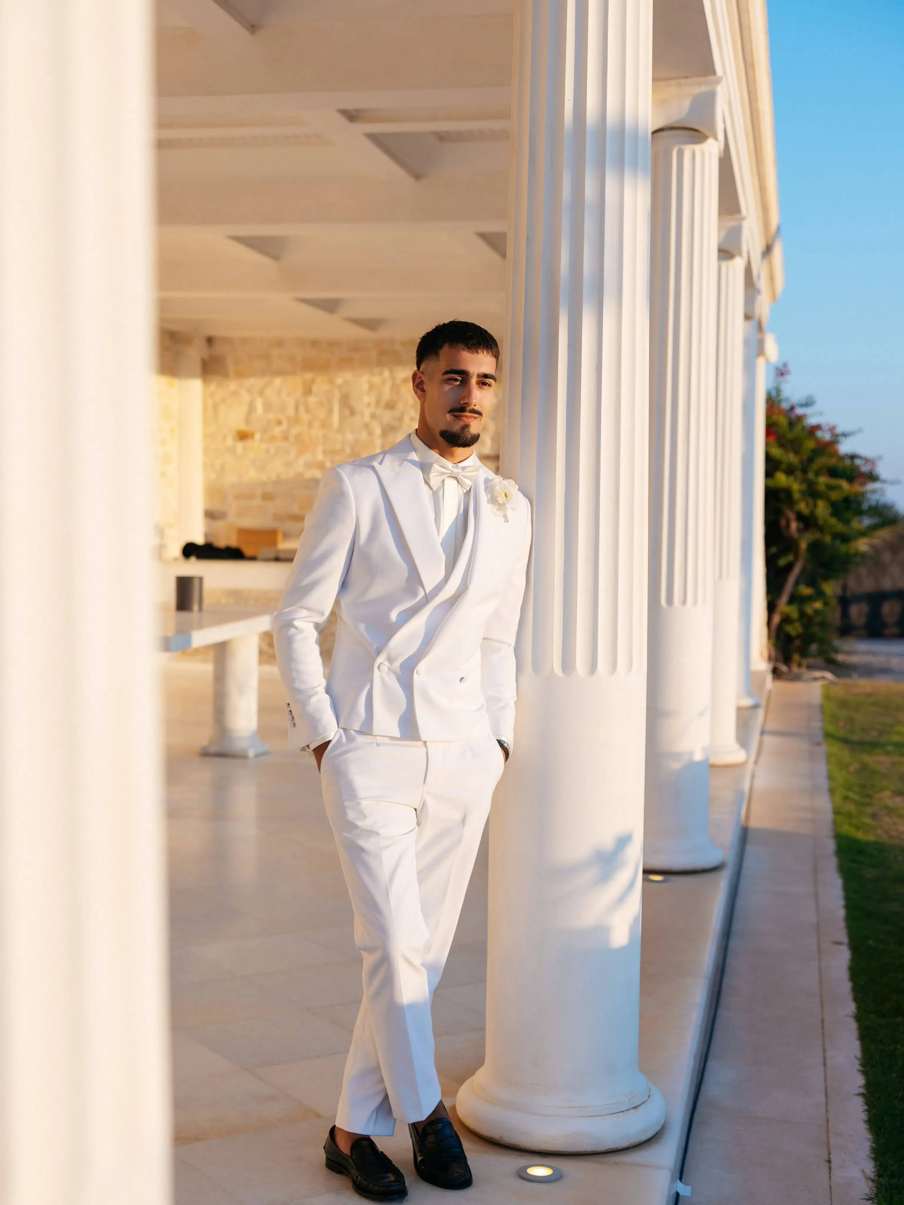 Groom in a white tuxedo leaning against a classical column at an outdoor venue