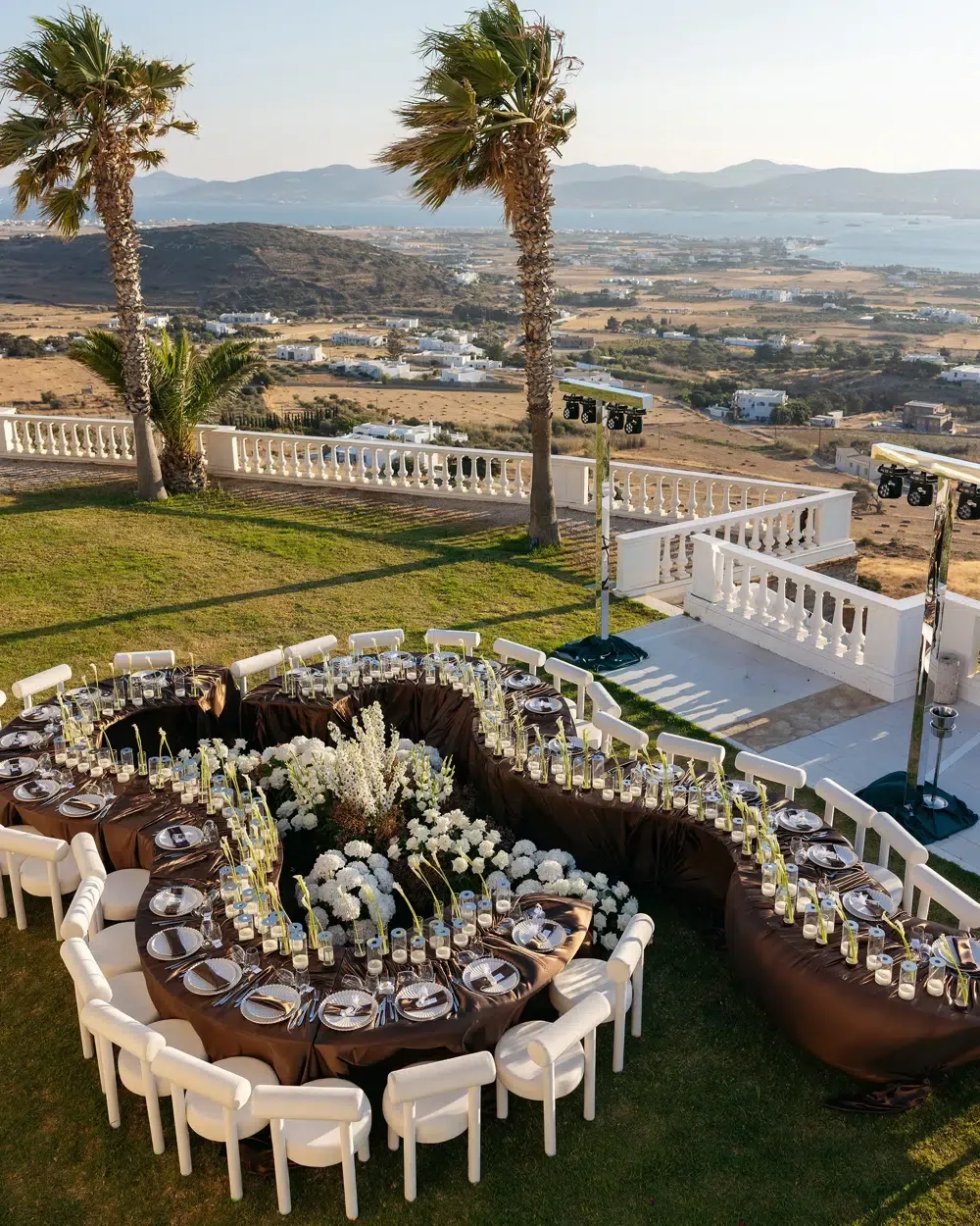 Aerial view of a winding serpentine wedding table lined with candles and white flowers on a coastal terrace