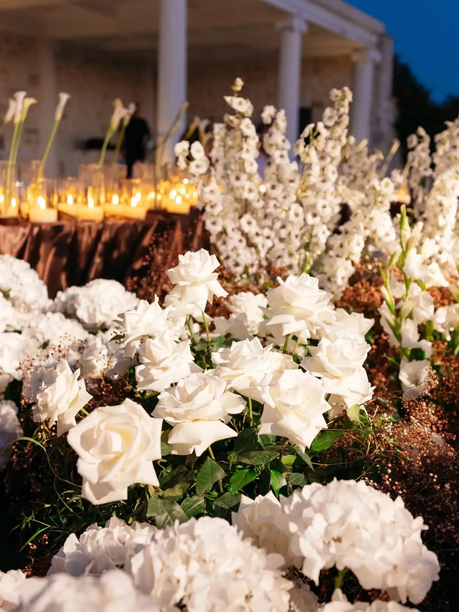 White roses and tall floral stems at an outdoor evening reception, with candlelight and classical columns behind
